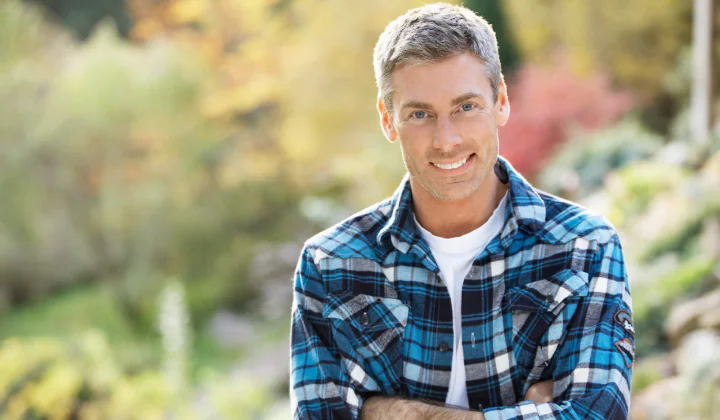 Older man smiling with crossed arms in a forest scene.